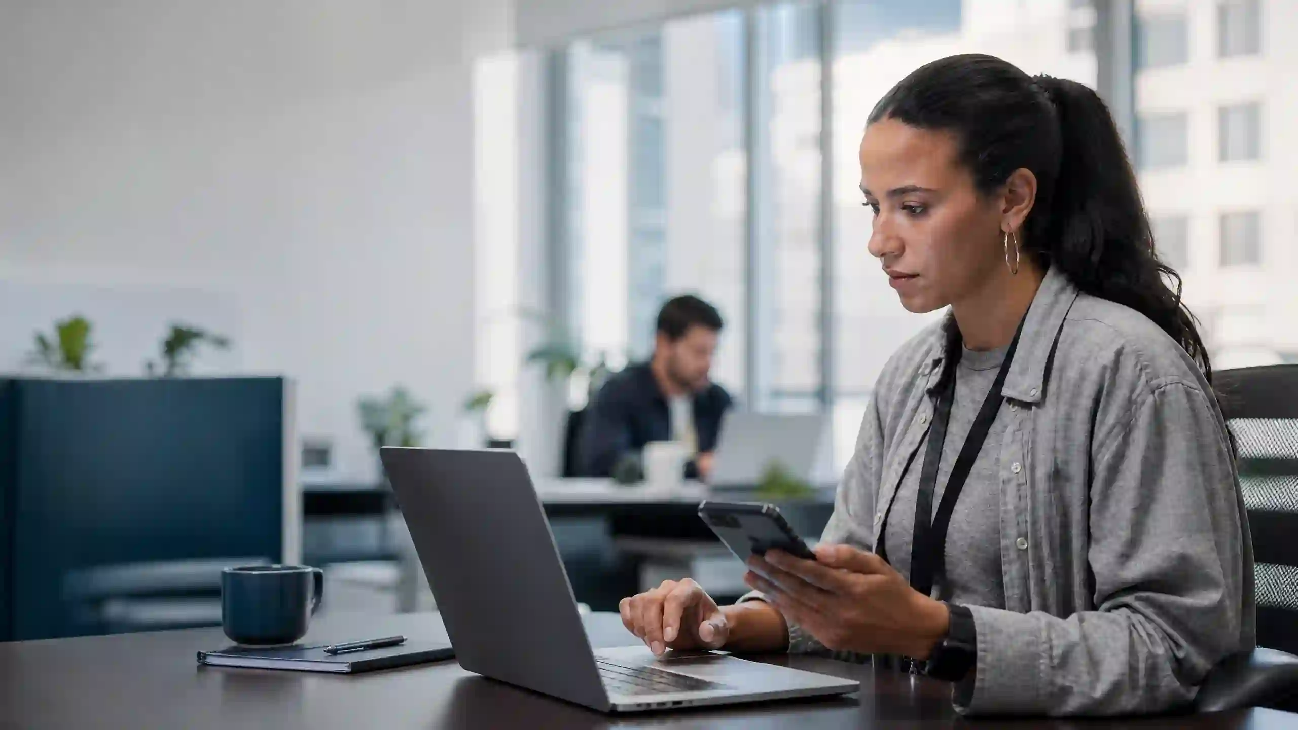 Profissional conferindo o celular ao lado do notebook em escritório, com colega trabalhando ao fundo